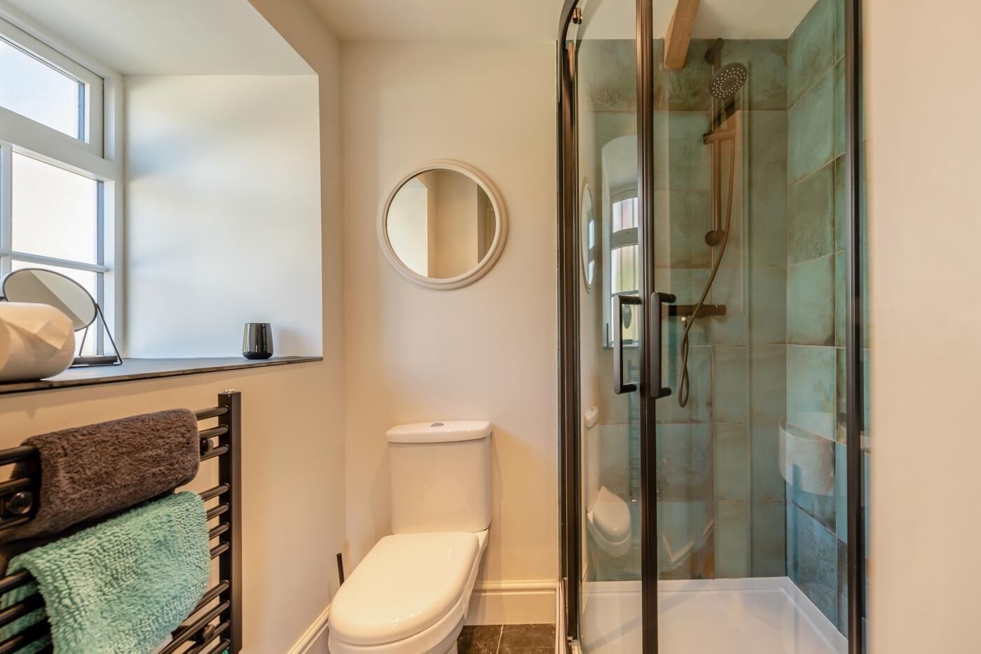 A bathroom with a toilet, a glass-enclosed shower, a round mirror, and a towel rack with towels.
