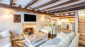 A cozy living room with a fireplace, wooden beams, a dining area, and a kitchen visible through an open door. - 4 Bed in Cartmel (oc-d31802) (Cartmel)
