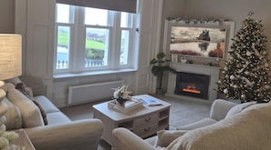 A cozy living room with a fireplace, a Christmas tree, and a large window with a view of the outdoors. - 2 Bed in Whitby (oc-e31738) (Whitby)