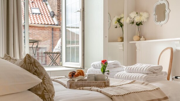 Cama bem arrumada com um tabuleiro de croissants e um vaso de flores, uma pilha de toalhas e vista para um prédio de tijolos pela janela.