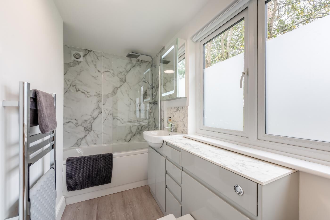 Een moderne badkamer met een douchewand van marmer, een badkuip, een wastafelmeubel met een grote spiegel, en een raam dat voor natuurlijk licht zorgt.