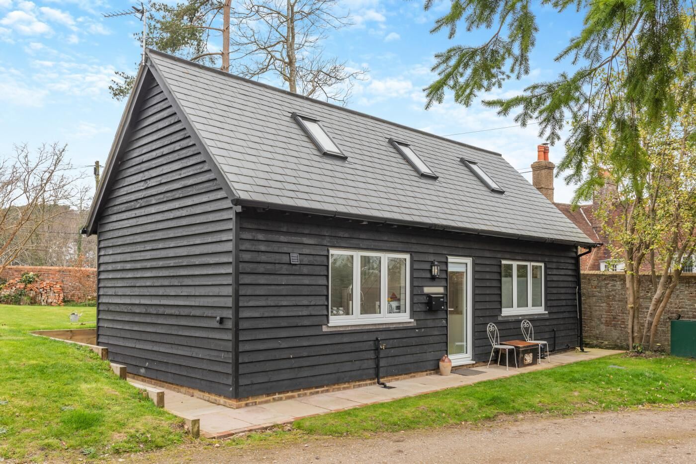 A small, dark-colored wooden house with a sloped roof, a front door, and a small patio area with a table and chairs.