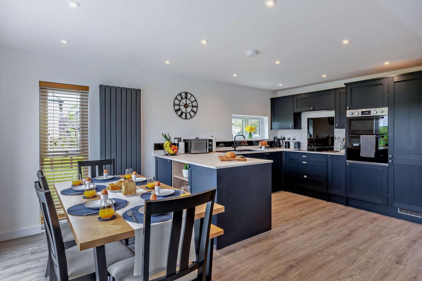 Een moderne keuken met donkere kastjes, een centraal eiland en een eethoek met een houten tafel en stoelen.