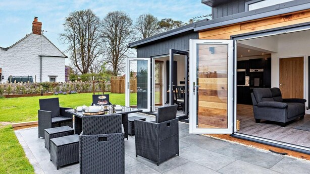 Una casa moderna con un patio amueblado y un ventanal corredero que da a un espacio interior con sofá y electrodomésticos de cocina.