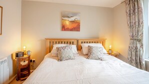 A double bed with a white quilt, two bedside tables with lamps, a painting above the bed, and a patterned curtain on the window. - 1 Bed in Llanfairfechan (oc-t31122) (Llanfairfechan)