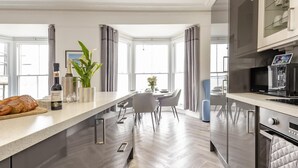 A modern kitchen with a long countertop, a dining area with chairs, and a view through large windows. - 2 Bed in Tenby (oc-o31794) (Tenby)