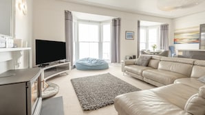 A modern living room with a flat-screen TV, a beige sofa, a blue bean bag, and a large window with curtains. - 2 Bed in Tenby (oc-o31794) (Tenby)