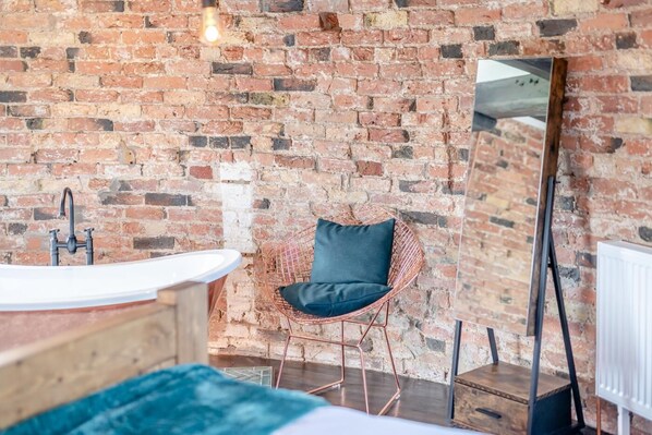 A bathroom with a brick wall, a round wire chair with a cushion, a white sink, and a mirror.