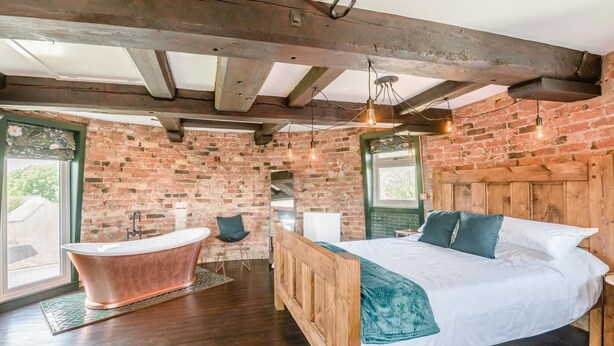 Ein Schlafzimmer mit einem hölzernen Bett, einem großen Fenster, einer Ziegelwand und einer Badewanne im Vintage-Stil.