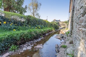 A narrow stream with a stone bridge, lush greenery, and a stone building on one side.