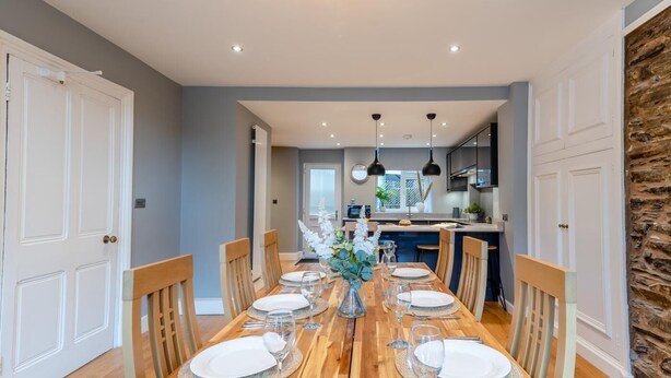 A dining room with a wooden table set for six, flanked by two doors and a kitchen area in the background. - 4 Bed in Windermere (oc-o31130) (Windermere)