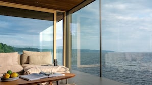 Ein modernes Zimmer mit großem Fenster mit Blick auf ein Gewässer, einem Sofa, einem Tisch mit Buch und Gläsern sowie einem Obstkorb.