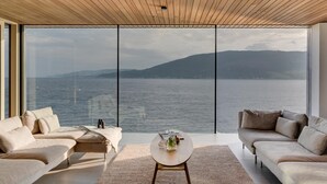 Ein modernes Wohnzimmer mit großem Fenster mit Blick auf ein Gewässer und Hügel. Im Zentrum befinden sich zwei Sofas und ein Couchtisch.