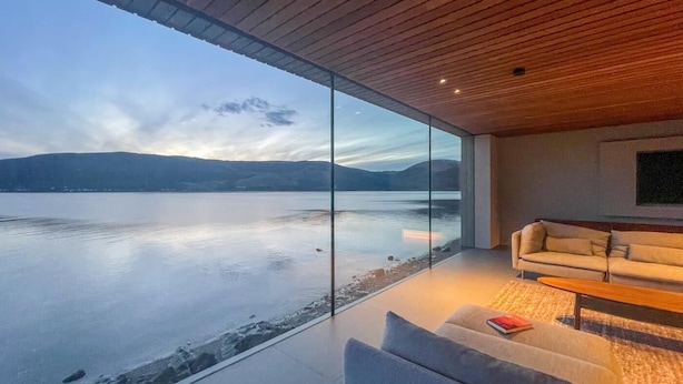 Ein modernes Wohnzimmer mit einer Glaswand mit Blick auf einen stillen See und Berge.