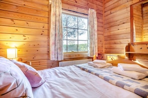A wooden cabin room with a bed, a window with curtains, a bedside table, and a radiator.