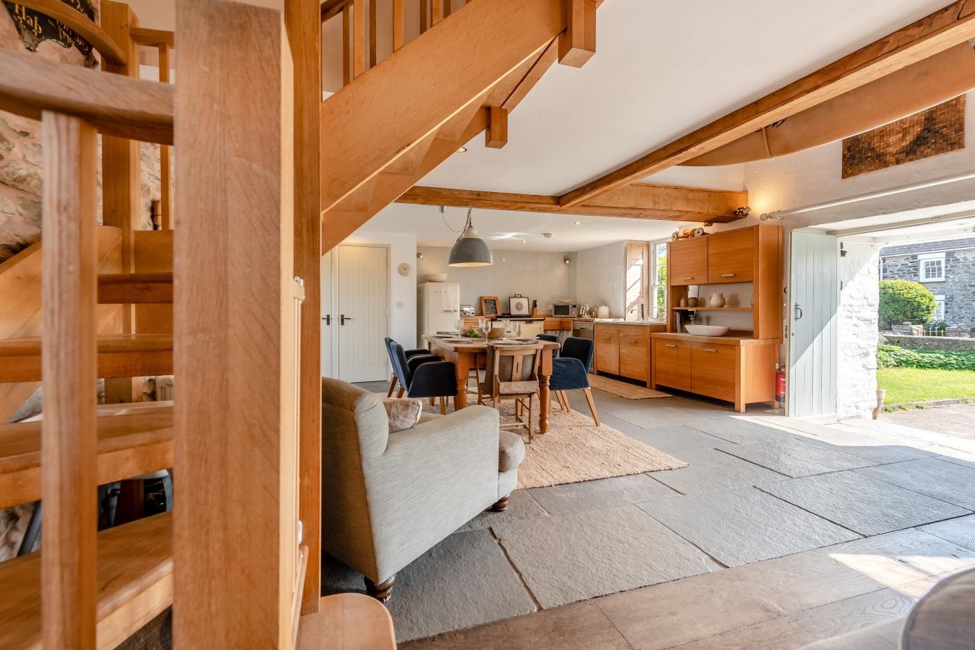 Een ruime woonkamer met een bank en stoelen, een eettafel, en een keuken met houten kasten en een stenen gevel.