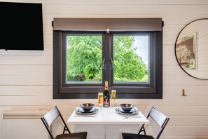 A dining area with a table set for two, a window with a view of greenery, and a mounted television on the wall.