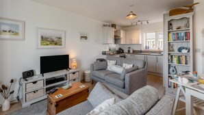 A modern living room with a grey sofa, a TV, and a coffee table. There is a kitchen in the background with white cabinets and a dining area with a table and chairs. - 2 Bed in Camber (oc-r31753) (Camber)