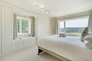 Ein Schlafzimmer mit einem großen Bett, zwei mit Vorhängen versehenen Fenstern, einem Nachttisch mit Lampe und Blick ins Grüne.