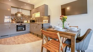 A modern kitchen with stainless steel appliances, a dining table set for four, and a large flat-screen TV mounted on the wall. - 2 Bed in Swanage (oc-s31851) (Swanage)