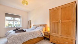 A bedroom with a bed, a wooden wardrobe, a bedside table with a lamp, and a window with a view of buildings. - 2 Bed in Swanage (oc-s31851) (Swanage)