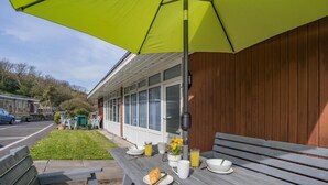 Zona de pati amb una banca de fusta, taula preparada amb plats, un got de suc i un para-sol groc.