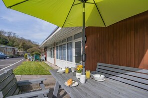 Un espace patio avec un banc en bois, une table dressée avec des assiettes, un verre de jus de fruits et un parasol jaune.