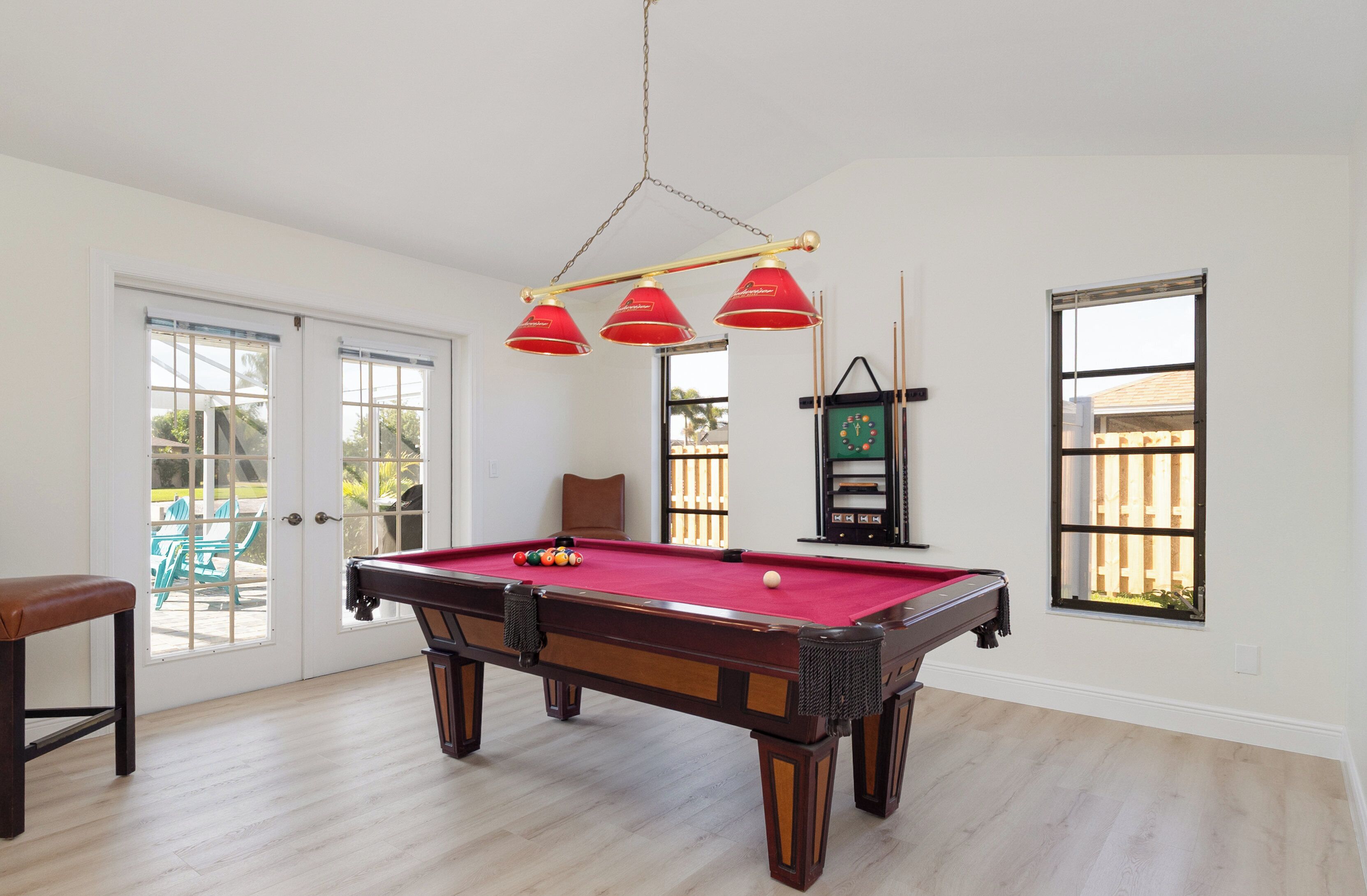 Una mesa de billar en una habitación con una puerta que da a un patio, un reloj en la pared y una ventana con vista a una cerca.