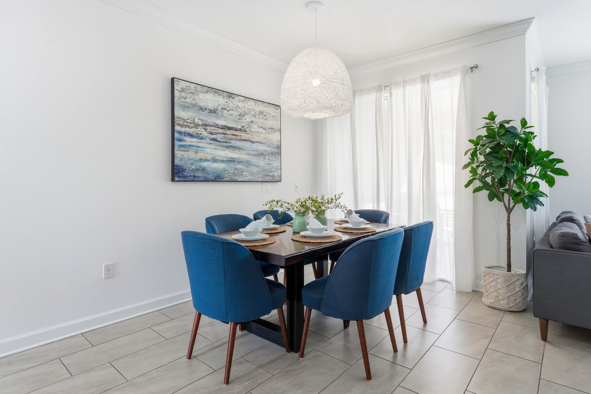 A dining area with a rectangular table set for four, blue upholstered chairs, a hanging light fixture, a painting on the wall, and a potted plant.