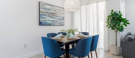A dining area with a rectangular table set for four, blue upholstered chairs, a hanging light fixture, a painting on the wall, and a potted plant.