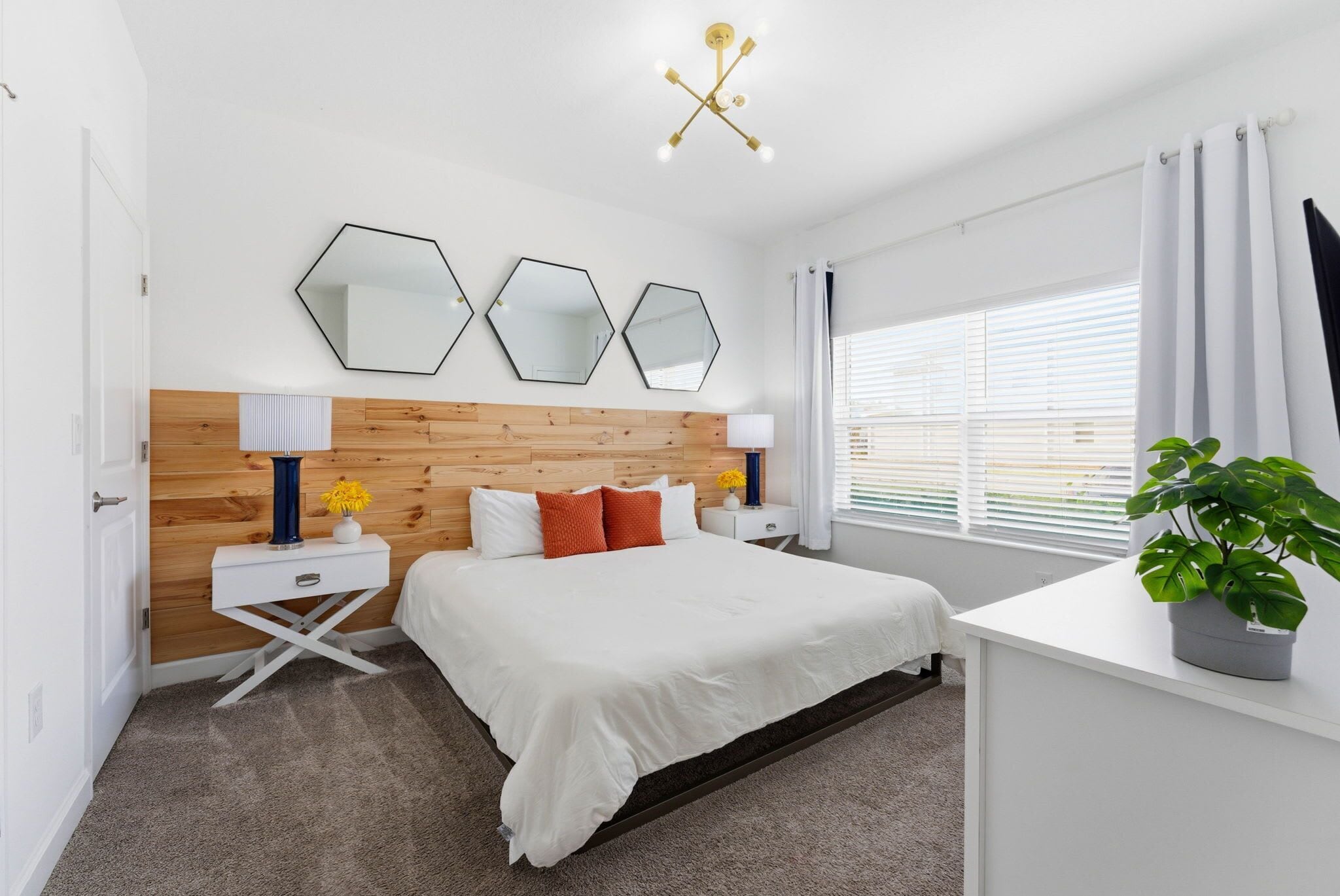 A modern bedroom with a wooden headboard, a large bed, a bedside table with a lamp, and a potted plant.
