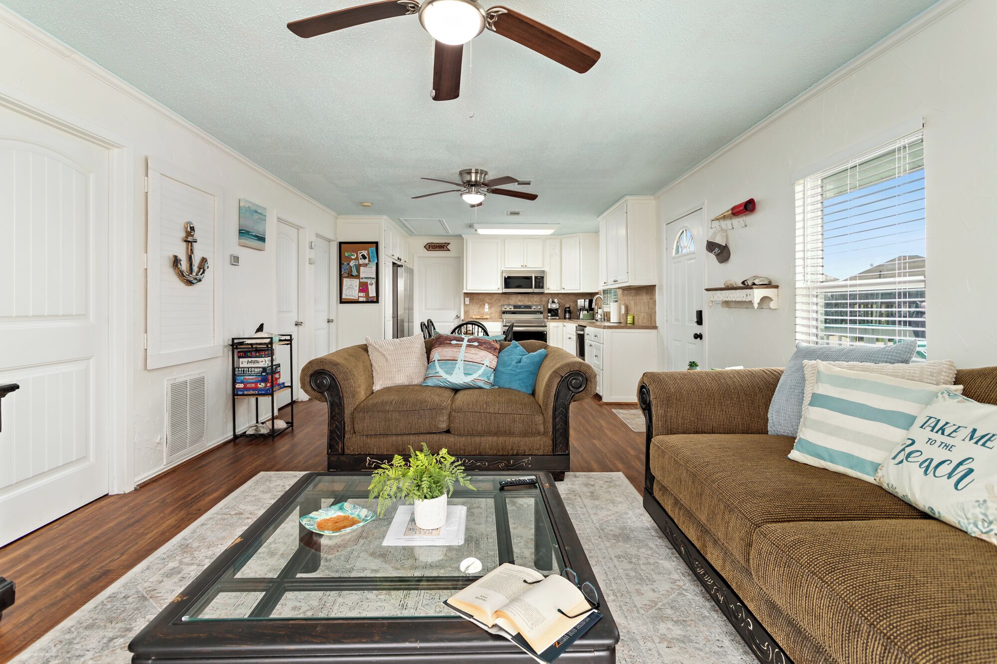 A living room with a glass coffee table, two brown sofas, a ceiling fan, and a kitchen in the background.