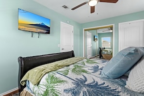 Ein Schlafzimmer mit einem Bett, einem Deckenventilator, einem an der Wand befestigten Fernseher und einer Tür zu einem anderen Raum.