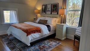 A neatly arranged bedroom with a large bed, a bedside table with a lamp, a window with curtains, and framed pictures on the wall. - Charming 1-bedroom apartment. Walk everywhere! A perfect Bellingham getaway. (Bellingham)