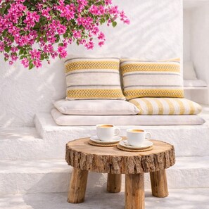 A round wooden table with two white cups and saucers, a white sofa with striped cushions, and a pink flowering plant. - Gloria Luxury Suites (Kassandra)