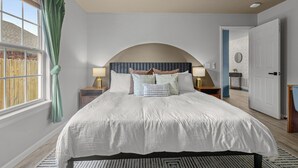 Une chambre à coucher avec un grand lit, deux tables de chevet avec des lampes, une fenêtre avec des rideaux et une porte donnant sur une autre pièce.