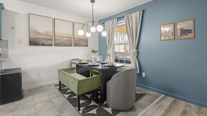 Un coin repas avec une table dressée pour quatre personnes, une chaise rembourrée de couleur verte et grise, une poubelle noire et un tapis à motifs.