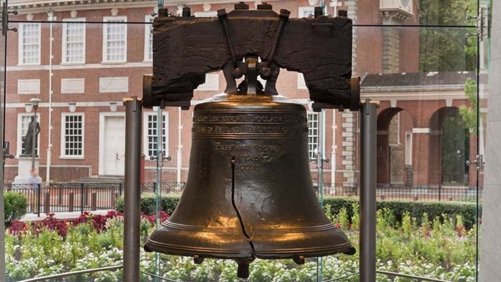 A large bell mounted on a metal frame with a plaque, set against a backdrop of a building with red brick walls and white windows.