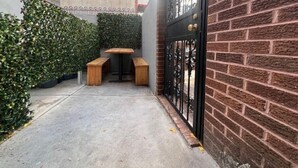 Un pequeño espacio al aire libre con una mesa de madera y bancos, rodeado por un muro de ladrillo y una reja metálica.