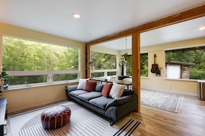 A spacious living area with a sofa, ottoman, and large windows offering a view of greenery.