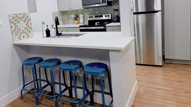 Una cocina moderna con armarios blancos, un refrigerador de acero inoxidable, un microondas y un lavamanos. Hay cuatro taburetes azules en la isla.