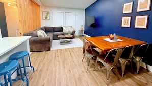 Una sala de estar moderna con una mesa de comedor de madera, taburetes de bar azules, un sofá gris y un televisor montado en la pared.