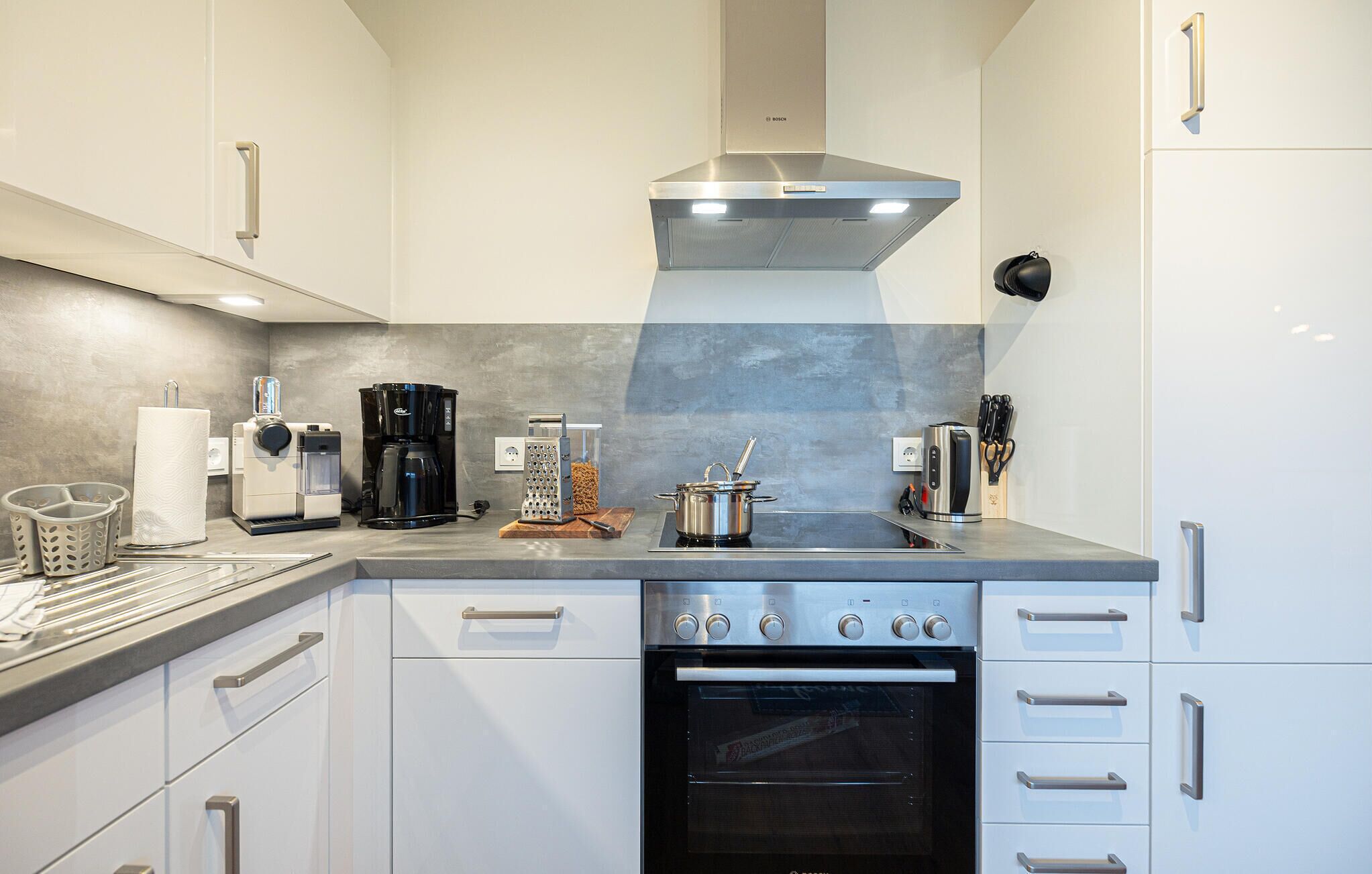 A modern kitchen with white cabinets, a Bosch range hood, a coffee maker, a kettle, a toaster, and a built-in oven.