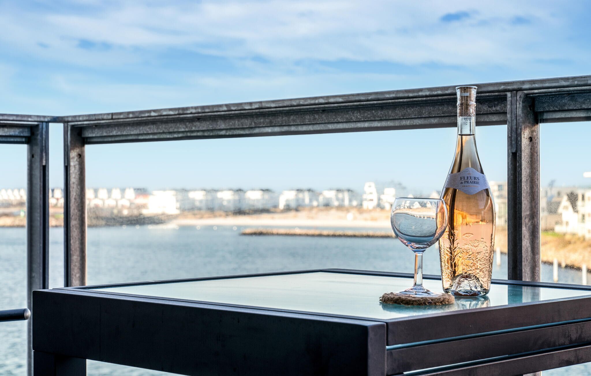 Une bouteille de vin et un verre sur une table, avec vue sur un pont et des bâtiments de l’autre côté d’une étendue d’eau.