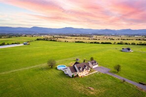 A large house with a swimming pool, surrounded by expansive green fields and mountains in the background.