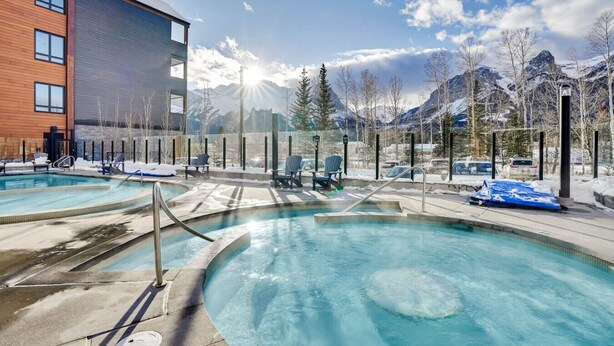 A pool area with multiple hot tubs and lounge chairs, surrounded by mountains and a building with large windows. - 3 Bedroom with Outdoor Heated Pool and Hottub! (Canmore)
