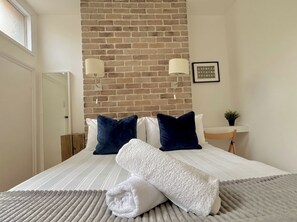 A hotel room with a brick wall, a bed with white and dark blue pillows, a white throw blanket, and a small table with a plant.