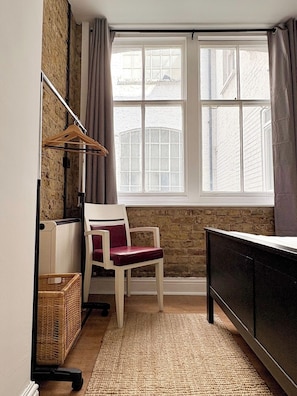 Ein Zimmer mit einem Bett, einem Sessel, einem Schreibtisch, einem Fenster und einem an der Wand befestigten Regal.