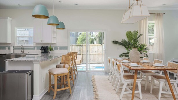 A modern kitchen with white cabinets, a granite countertop, and pendant lighting. There is a dining area with a wooden table and chairs, and a large plant in the center. - Family Beach Escape w/ Private Pool|Downtown St. A (St. Augustine)