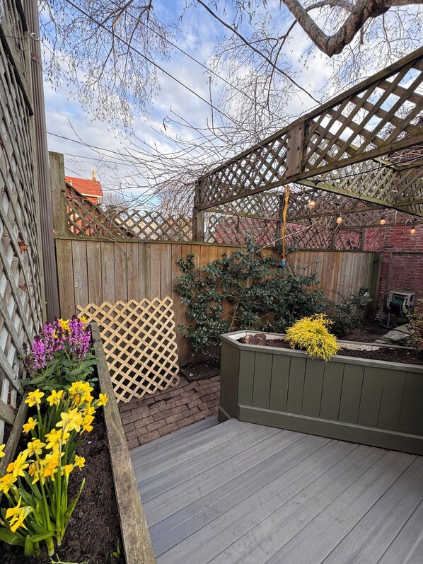 A wooden deck with a lattice structure overhead, surrounded by a wooden fence and a brick wall.
